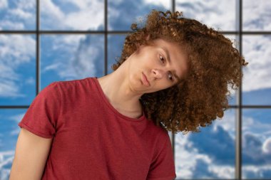 Koyu kırmızı tişörtlü genç adam kıvırcık saçlarıyla boğuşuyor, stüdyoda duruyor, fotokopi çekiyor. Kıvırcık saçlarını sallayan bir adam. Kuru ve mat saçlar.