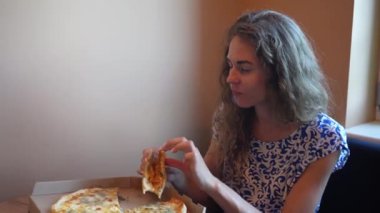 Genç bir kadın pencerenin yanında oturuyor ve taze teslim edilmiş bir pizza yemek üzere. O, beyaz ve mavi bir elbise giyiyor, doğal ışık ile sıcak bir odada yemeğinin tadını çıkarıyor.