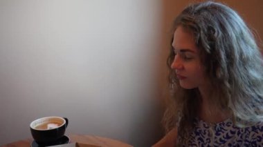 Mavi ve beyaz elbiseli genç bir kadının yakın plan fotoğrafı bir masada oturuyor, içeride bir fincan kahvenin tadını çıkarıyor. Yumuşak doğal ışık, o anın huzurlu ve rahat atmosferini vurgular.