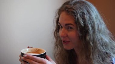 Mavi ve beyaz elbiseli genç bir kadının yakın plan fotoğrafı bir masada oturuyor, içeride bir fincan kahvenin tadını çıkarıyor. Yumuşak doğal ışık, o anın huzurlu ve rahat atmosferini vurgular.