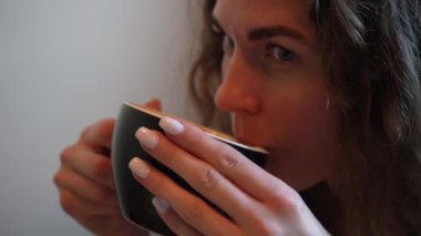 Yumuşak doğal ışık, o anın sakin ve samimi atmosferini arttırır. Mavi beyaz elbiseli genç bir kadın, kapalı bir masada otururken bir yudum kahve alır.