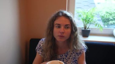 Mavi ve beyaz elbiseli genç bir kadının yakın plan fotoğrafı bir masada oturuyor, içeride bir fincan kahvenin tadını çıkarıyor. Yumuşak doğal ışık, o anın huzurlu ve rahat atmosferini vurgular.