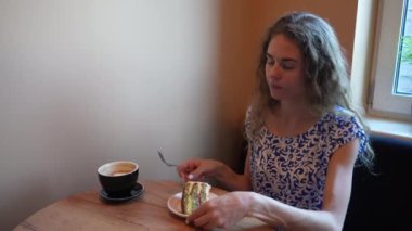 Genç bir kadın pencerenin kenarındaki rahat bir masada oturmuş, rahat, doğal bir ortamda kahvesinin tadını çıkarmaya hazır bir dilim pastanın tadını çıkarıyor.