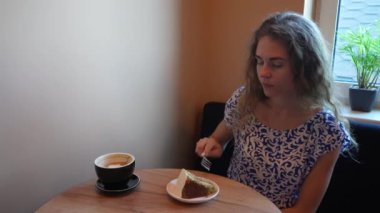 Genç bir kadın pencerenin kenarındaki rahat bir masada oturmuş, rahat, doğal bir ortamda kahvesinin tadını çıkarmaya hazır bir dilim pastanın tadını çıkarıyor.