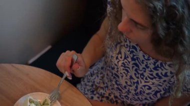 Mavi ve beyaz elbiseli bir kadın ahşap bir masada oturur, sıcak bir ortamda kahvesini yudumlayarak bir dilim pastanın tadını çıkarır, yemeğini yemeye hazırdır.