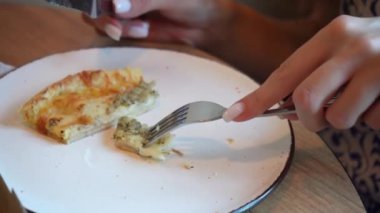 Genç bir kadın bir dilim pizzayı tadıyor. Bir dilim pizza beyaz bir tabakta duruyor, kız da çatal bıçakla kesiyor.