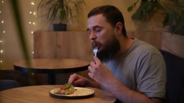 Bir adam tabağına konsantre olur, dikkatle bir waffle ve sebzeleri dilimler. Kafedeki hafif ışıklandırmayla yemeğin tadını çıkarıyor.