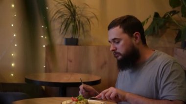 Bir adam tabağına konsantre olur, dikkatle bir waffle ve sebzeleri dilimler. Kafedeki hafif ışıklandırmayla yemeğin tadını çıkarıyor.