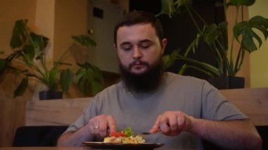 Çatal ve bıçak çatalı kullanan bir adam yemek siparişini tüketir. Genç sakallı bir adam bir kafe masasında oturup sıcak ve yumuşak bir ortamda yemeğin tadını çıkarıyor.