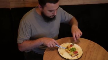 Çatal ve bıçak çatalı kullanan bir adam yemek siparişini tüketir. Genç sakallı bir adam bir kafe masasında oturup sıcak ve yumuşak bir ortamda yemeğin tadını çıkarıyor.