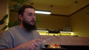 Genç sakallı bir adam bir kafe masasında oturup sıcak ve yumuşak bir ortamda yemeğin tadını çıkarıyor. Çatal ve bıçak çatalı kullanan bir adam yemek siparişini tüketir.