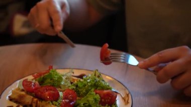 Yakından, çatal ve bıçakla taze domates salatası yiyen bir adam. Tabak, rahat bir atmosferde sağlıklı yiyecekleri ön plana çıkaran ahşap bir masada servis ediliyor.