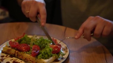 Yakından, çatal ve bıçakla taze domates salatası yiyen bir adam. Tabak, rahat bir atmosferde sağlıklı yiyecekleri ön plana çıkaran ahşap bir masada servis ediliyor.