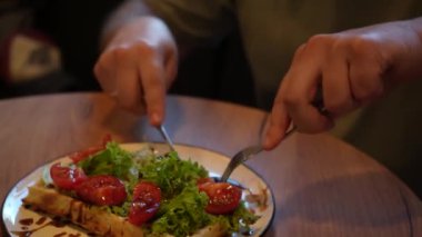 Yakından, çatal ve bıçakla taze domates salatası yiyen bir adam. Tabak, rahat bir atmosferde sağlıklı yiyecekleri ön plana çıkaran ahşap bir masada servis ediliyor.