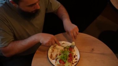 Sakin atmosfer yiyeceğin rahatlama ve keyif alma anını vurgular. Genç sakallı bir adam bir kafeteryanın arka planında oturuyor, sıcaklığın tadını çıkarıyor.