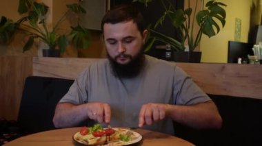 Loş bir kafede, ahşap bir masada yemek yiyen sakallı bir adamın yakın çekimi. Yumuşak atmosfer ve erkeğin yemeklerine yoğunlaşması, tek başına huzurlu bir yemek anının basitliğini vurgular.