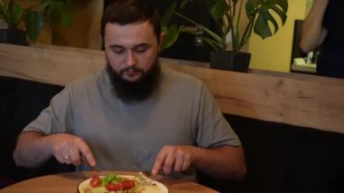 Sıcak bir ışık ve sakin bir atmosfer onu sarar. Uzun bir günün ardından gevşemek için mükemmel bir yemek deneyimi yaratır. Sakallı bir adam yemeğin tadını çıkarır, yemek tabağına odaklanır..