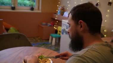 Sakallı bir adam yemek yemekten zevk alır, yemek tabağına odaklanır. Sıcak bir ışık ve sakin bir atmosfer etrafını sarar. Uzun bir günün ardından gevşemek için mükemmel bir yemek deneyimi yaratır.