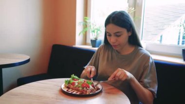 Bir kadın pencerenin kenarında oturmuş, rahat bir evde sağlıklı bir yemeğin tadını çıkarıyor. Sahne, huzuru, farkındalığı ve temiz yemeğe odaklanmayı ifade ediyor.