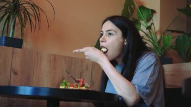 Pencerenin yanındaki küçük bir masada taze sebze yemeğinin tadını çıkaran genç bir kadın. Doğal ışık yemek deneyiminin basitliğini ve sakinliğini arttırır.