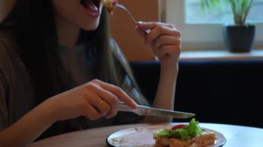 Gündelik giyinmiş bir kadın yemeğini düşünceli bir şekilde ev ortamında yer. Parlak gün ışığı penceresinden süzülüp sağlıklı yiyeceklerinin tazeliğini vurguluyor.