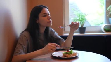 Gündelik giyinmiş bir kadın yemeğini düşünceli bir şekilde ev ortamında yer. Parlak gün ışığı penceresinden süzülüp sağlıklı yiyeceklerinin tazeliğini vurguluyor.