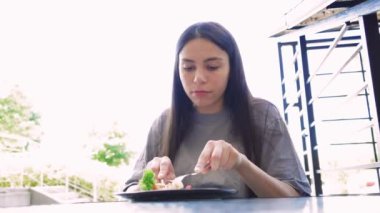 Arka planda doğal ışık ve yeşillik sakin ve taze bir atmosfer yaratır. Dışarıda sağlıklı bir yemeğin tadını çıkaran genç bir kadın çatal ve bıçakla yemek yerken tabağına odaklanmış.