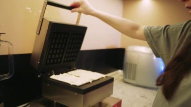 Genç bir kadın elektrikli waffle makinesine hamur dökerek lezzetli sonuçları sabırsızlıkla bekliyor. Taze pişmiş waffle 'ların tatlı kokusu mutfağı dolduruyor ve enfes bir atmosfer yaratıyor.