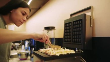 Genç bir kadın elektrikli waffle makinesine hamur dökerek lezzetli sonuçları sabırsızlıkla bekliyor. Taze pişmiş waffle 'ların tatlı kokusu mutfağı dolduruyor ve enfes bir atmosfer yaratıyor.