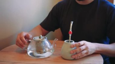 Bir adamın beyaz seramik bardaktan metal bir pipetle çay içtiği sıradan bir an. Arka planda demlik buharı