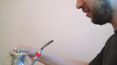Rahat bir adam, metal bir pipetle eşsiz bir bardaktan su içiyor, cam bir çaydanlığın yanında oturuyor. Sakin, rahat atmosfer