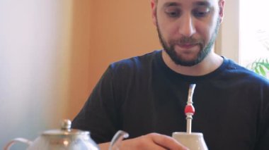 Bir çaydanlık masada buharlaşıyor, atmosfer sakin ve rahat. Bir adam geleneksel seramik bardaktan çay içer, derin düşüncelere dalmış.