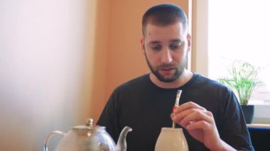 Geleneksel seramik bardaktan çay yudumlayan bir adam, derin düşüncelere dalmış. Çaydanlık buharlaşıyor, atmosfer sakin ve rahat.