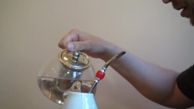 Geleneksel seramik bardaktan çay yudumlayan bir adam, derin düşüncelere dalmış. Çaydanlık buharlaşıyor, atmosfer sakin ve rahat.