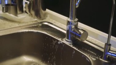Mutfakta bulaşık yıkayan biri, akan suyun altında parlak metal bir lavaboyu temizler. Resim günlük ev ödevlerini iş başında gösteriyor
