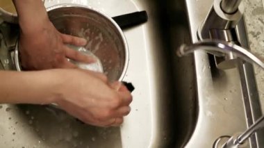 Lavaboda akan suyun altında paslanmaz çelik bir kase yıkayan ellerin yakın çekimi. Gündelik mutfak rutinine ve temizlik sürecine odaklanıyoruz.