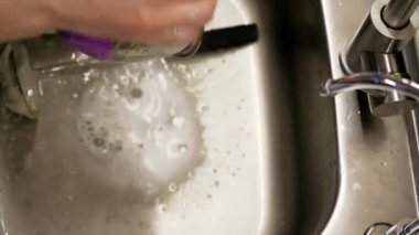 Lavaboda akan suyun altında paslanmaz çelik bir kase yıkayan ellerin yakın çekimi. Gündelik mutfak rutinine ve temizlik sürecine odaklanıyoruz.