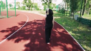 Şehir hayatı ve sessiz düşüncenin bir kombinasyonu. Genç bir kadın elinde bir fincan çay tutarken özenle arka planda duran spor sahası olan bir konut binasının önünde duruyor.
