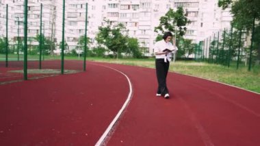 Elinde bir kitapla kırmızı bir koşu bandında, konut binalarının yakınındaki bir parkta yürüyen bir kadın. Şehir hayatı ve açık hava temaları için harika