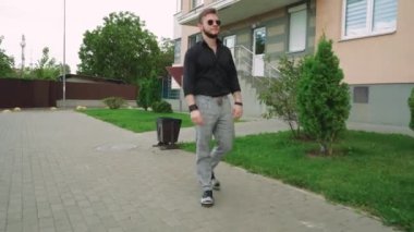 Güneş gözlüklü bir adam yeşil bir parkta bir şehir tarafından çevrili bir fayans yolu boyunca yürür. Rahatlama, yalnızlık ve açık hava yaşam tarzı temaları için mükemmel.