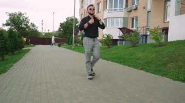 Güneş gözlüklü bir adam yeşil bir parkta bir şehir tarafından çevrili bir fayans yolu boyunca yürür. Rahatlama, yalnızlık ve açık hava yaşam tarzı temaları için mükemmel.