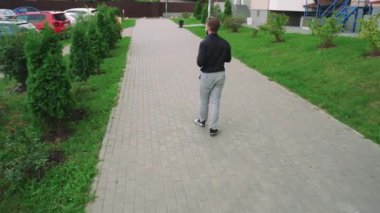 Siyah tişörtlü ve gri kotlu vahşi bir adam kaldırım boyunca yürür konut binalarının arka planında