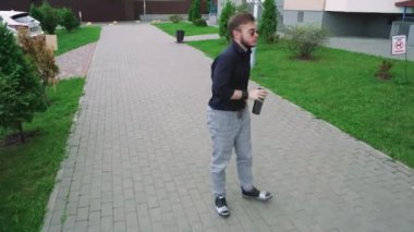 Güneş gözlüklü ve elinde bir fincan çay olan bir adam konut binalarının yakınındaki yeşil bir parktan geçiyor. Rahatlama, yalnızlık ve açık hava yaşam tarzı temaları için mükemmel.