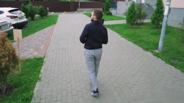 Güneş gözlüklü ve elinde bir fincan çay olan bir adam konut binalarının yakınındaki yeşil bir parktan geçiyor. Rahatlama, yalnızlık ve açık hava yaşam tarzı temaları için mükemmel.