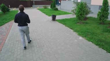 Güneş gözlüklü bir adam yeşil bir parkta bir şehir tarafından çevrili bir fayans yolu boyunca yürür. Rahatlama, yalnızlık ve açık hava yaşam tarzı temaları için mükemmel.