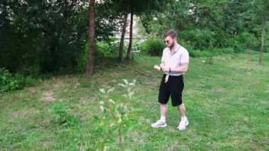 Güneş gözlüklü genç bir adam bitki ve ağaçlarla çevrili çimenli bir alanda sakince geziniyor. Açık hava içeriği için ideal