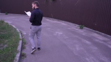 Bir adam, elinde bir kitapla, kahverengi metal bir çitin arkasında huzurlu bir şekilde yürümekten hoşlanır. Bu huzurlu sahne görsel rahatlama, huzur ve doğa keşfi içindir.