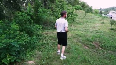 Genç adam yemyeşil bir park alanında yürüyor, yemyeşil bitkiler ve ağaçlarla çevrili. Yaşam tarzı ve açık hava rahatlama temaları için mükemmel