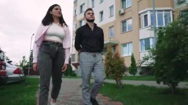 Bir kadın ve genç bir adam, sıradan giysiler içinde, kolları omzuna sarılı bir şekilde bir yerleşim tesisinin yakınlarında dolaşarak, şehirde rahat bir atmosfer yaratıyor.