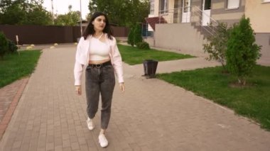 Sıradan giyinmiş, ceketli bir kadın yeşil kentsel alanda yürüyor. Etrafı apartmanlarla çevrili. Modern yaşam tarzı ve kentsel imajlar için uygun